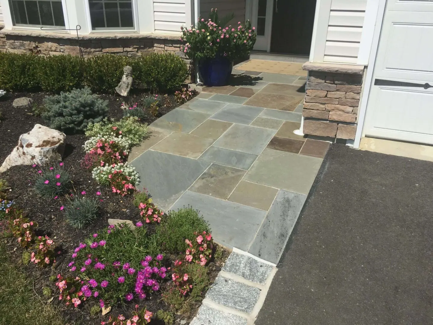 A stone walkway leading to a house entrance with surrounding flowers and greenery.