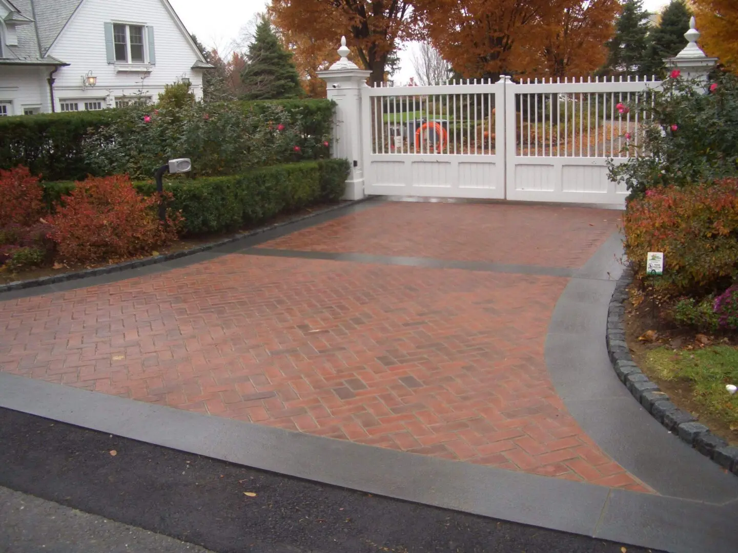 Brick driveway leading to a white gate with autumn foliage.