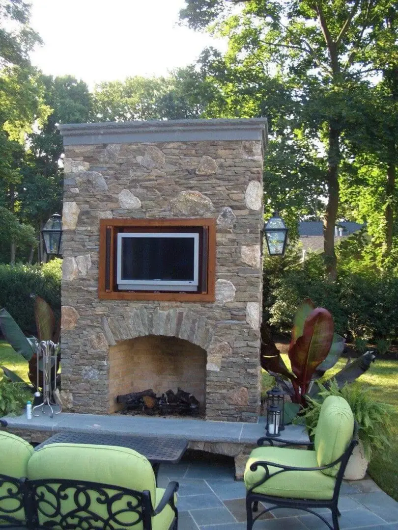 Outdoor stone fireplace with mounted TV in a lush backyard.