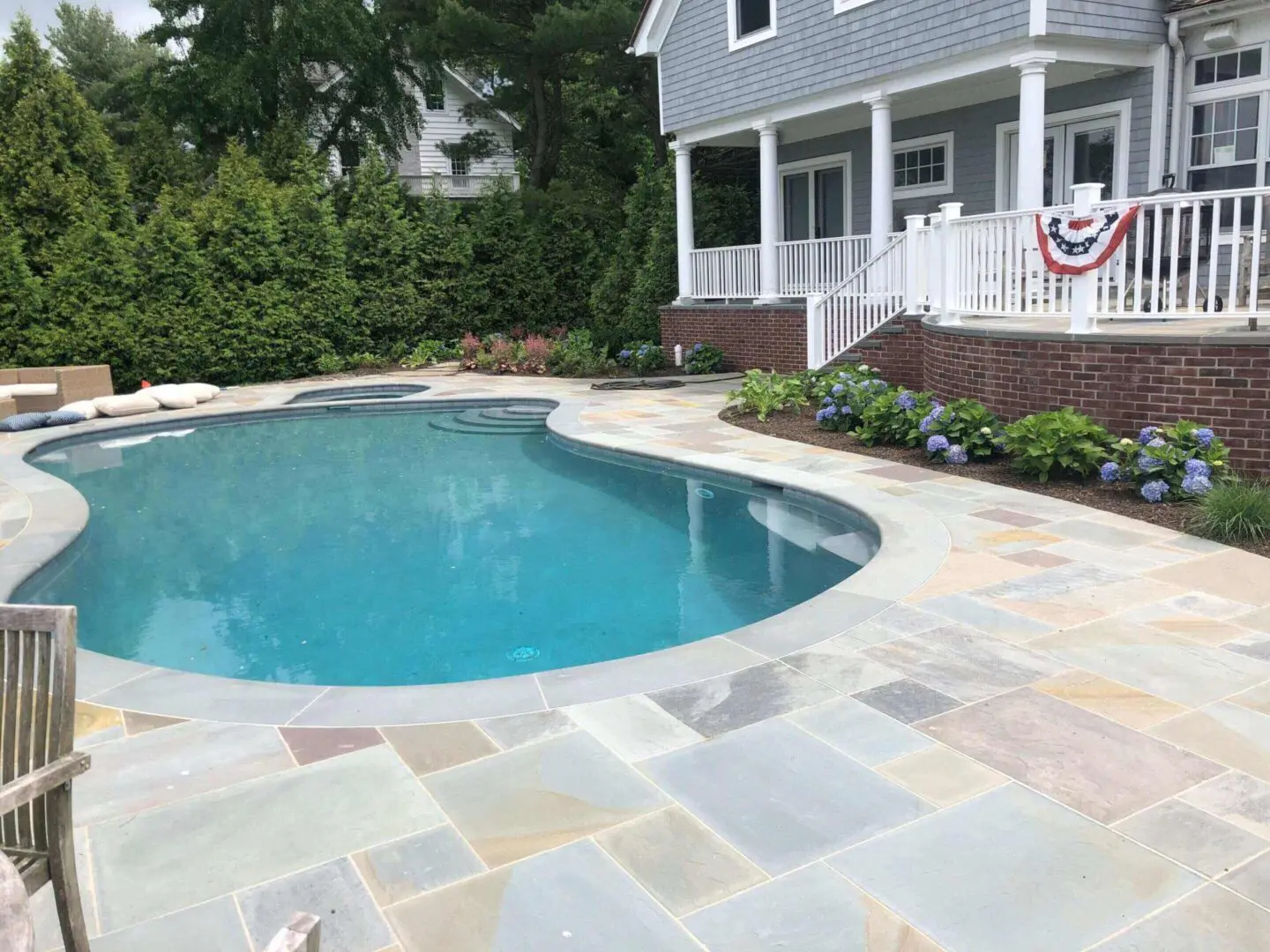 A clean, curved backyard pool with surrounding stone patio and lush greenery.