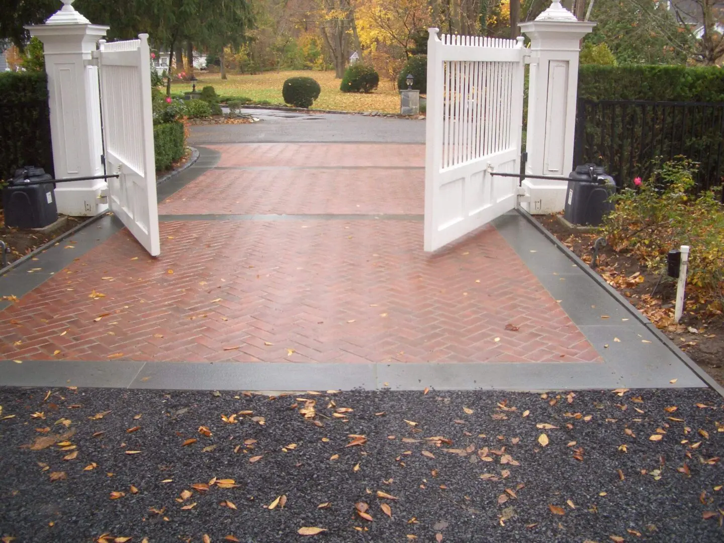 Open white gates leading to a brick driveway with autumn leaves scattered.