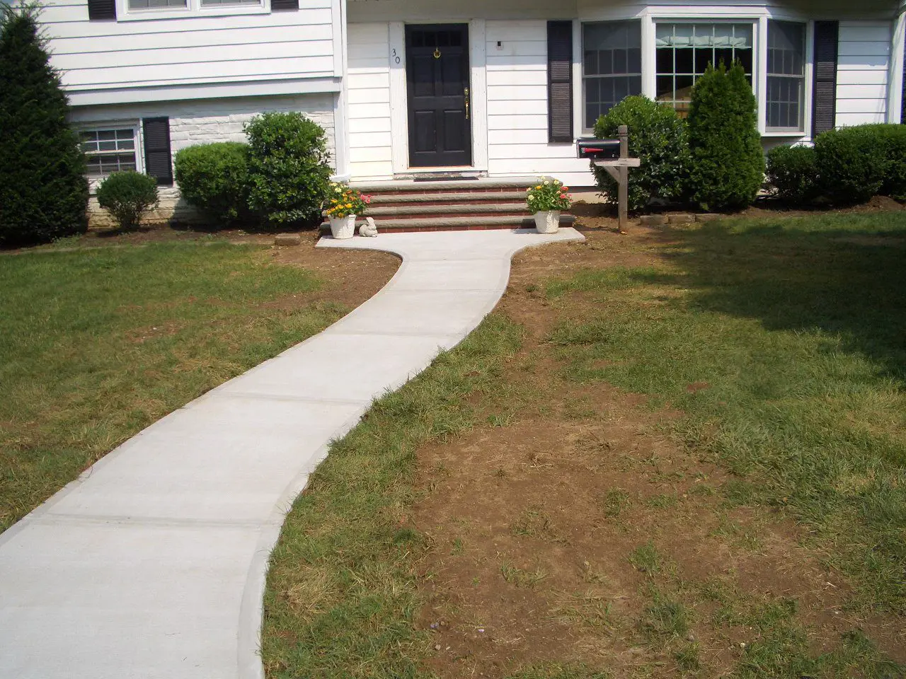 Concrete walkway leading to a house entrance with patchy grass on either side.