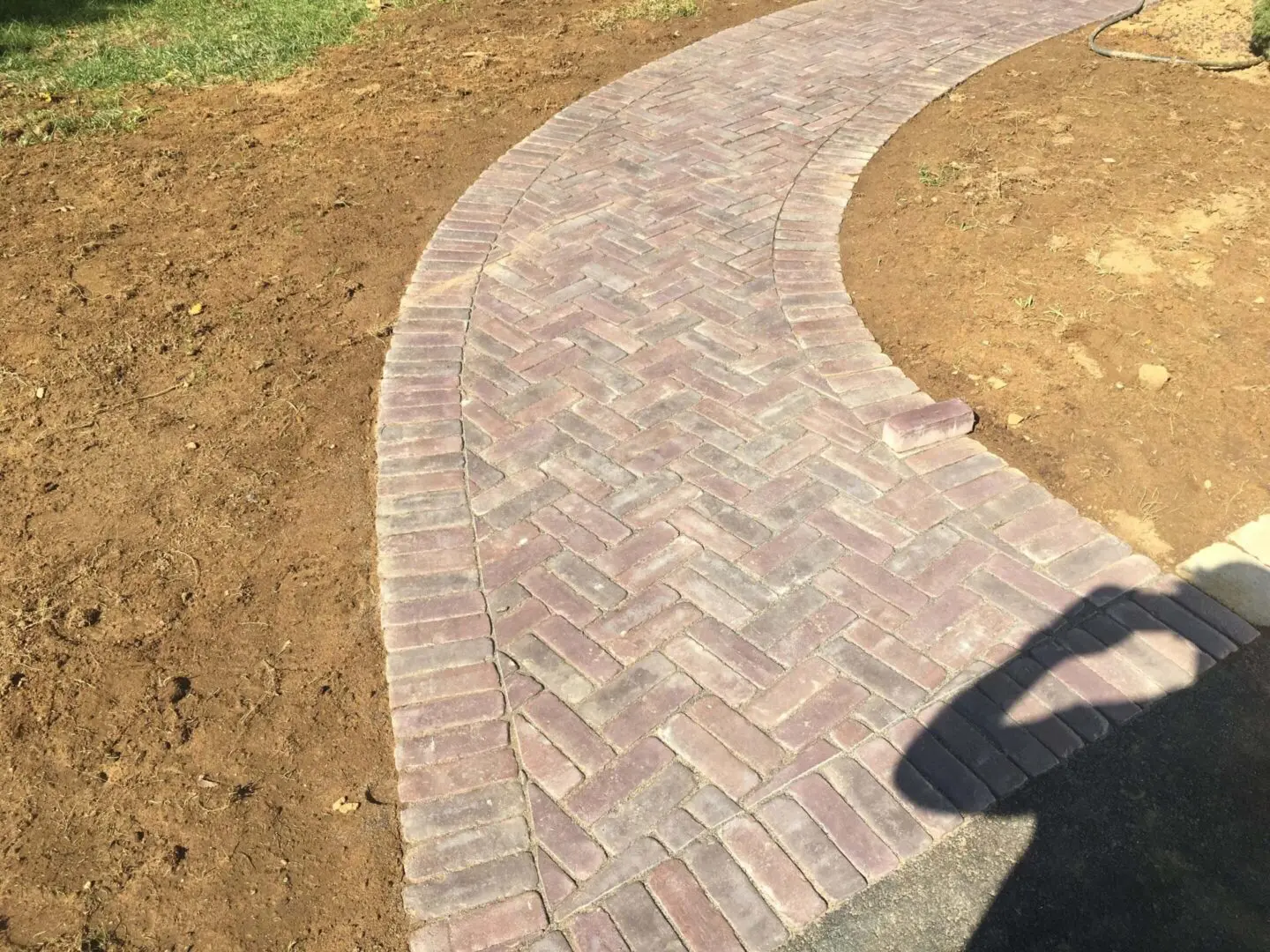 Curved brick pathway bordered by dry soil and a shadow.